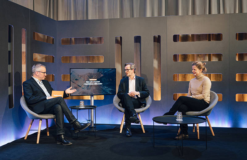 Tre personer sitter i en studio, engagerade i en diskussion framför en skärm, med modern belysning och minimalistisk design.